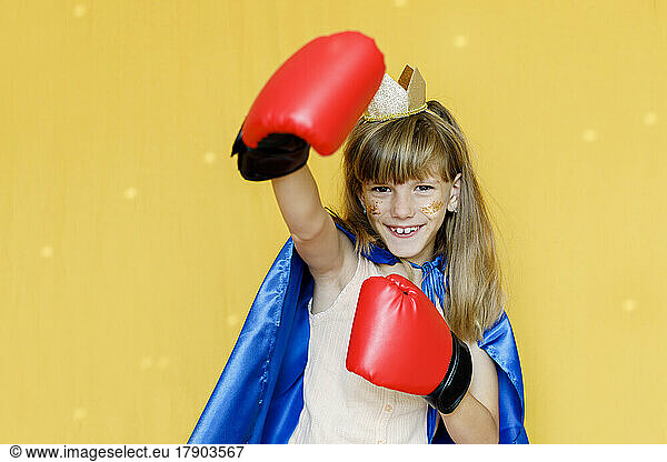 Happy girl wearing boxing gloves and cape against yellow background