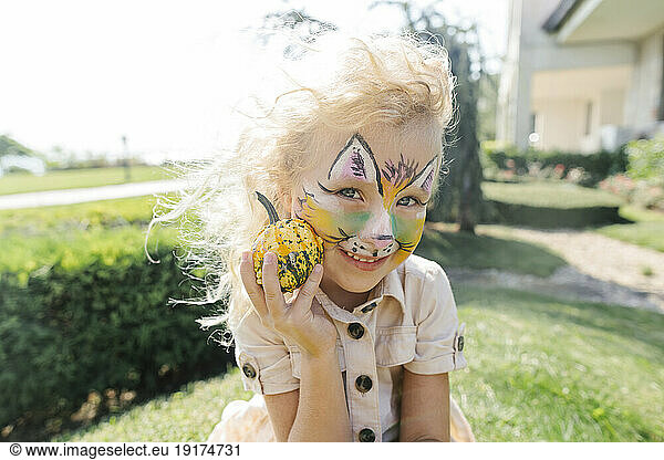 Happy girl with face painting holding small pumpkin on sunny day