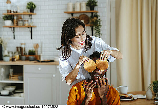Happy girlfriend holding pancakes in front of boyfriend's eyes in kitchen