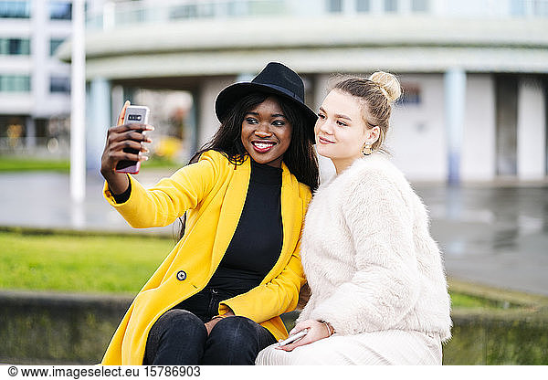 Happy girlfriends sitting on a bench in the city taking a selfie