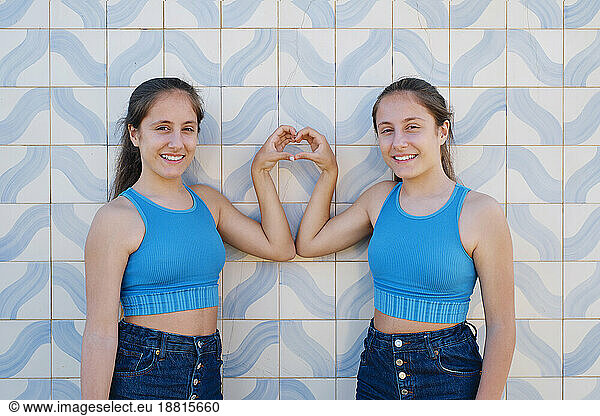 Happy girls gesturing heart shape in front of wall