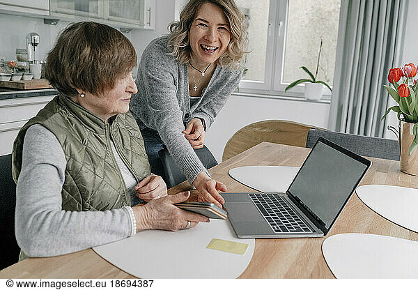 Happy granddaughter by senior woman with laptop on table at home