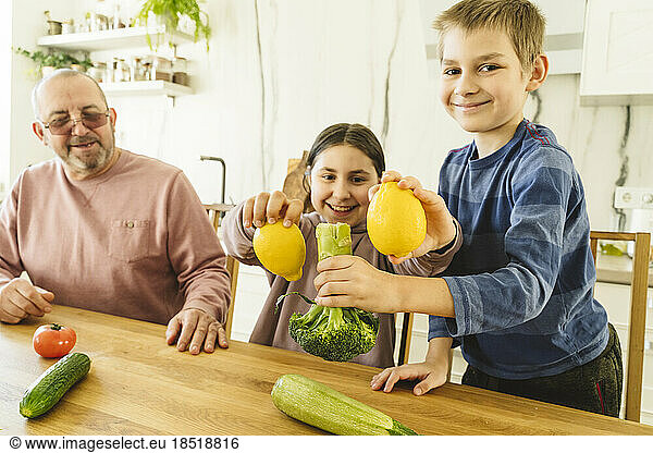 Happy grandfather with grandson and granddaughter holding vegetables at dining table