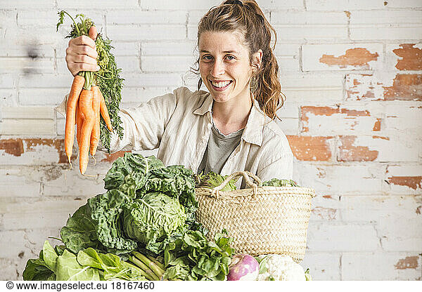 Happy grocer holding bunch of carrots in front of brick wall