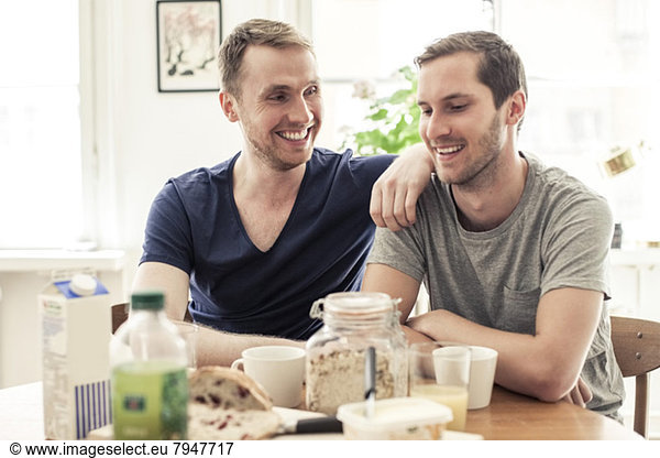 Happy homosexual couple communicating while having breakfast together at table in home