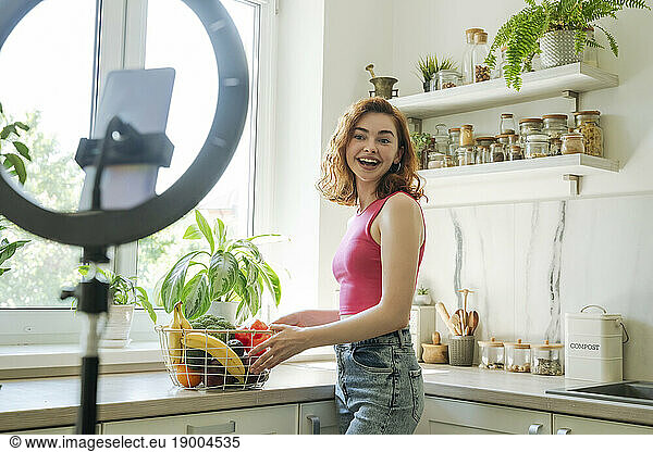 Happy influencer with basket of fruits and vegetables standing by kitchen counter at home