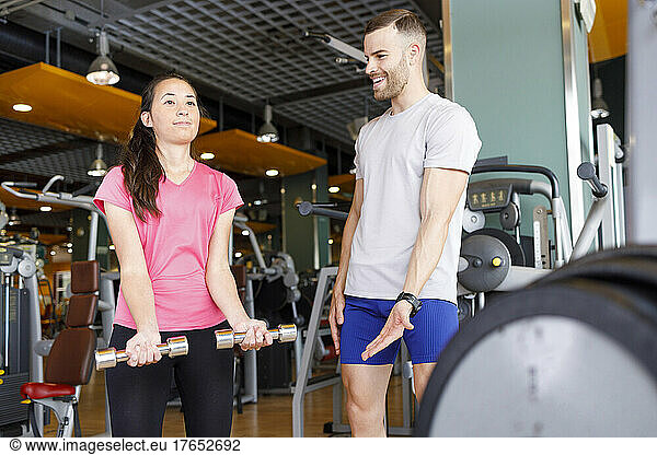 Happy instructor motivating woman exercising with dumbbells at gym