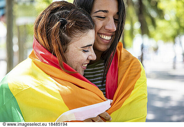 Happy lesbian couple covered in rainbow colored flag on footpath