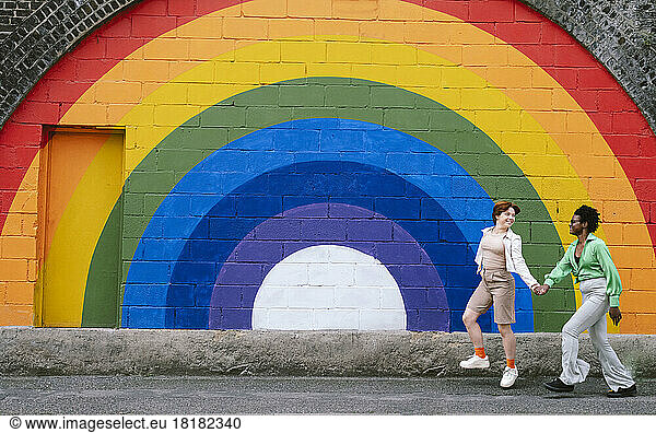 Happy lesbian couple holding hands walking on footpath in front of rainbow colored wall