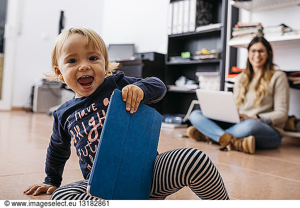 Happy little girl sitting on the floor at home with tablet and mother in background