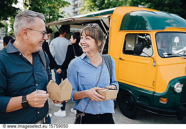Happy male and female friends talking while standing with box against commercial land vehicle