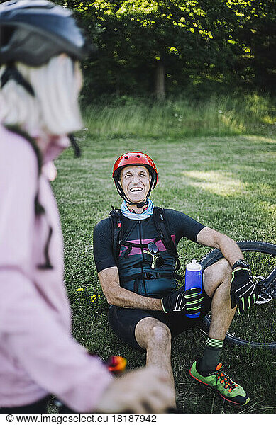 Happy male cyclist sitting on grass holding water bottle while talking to female friend