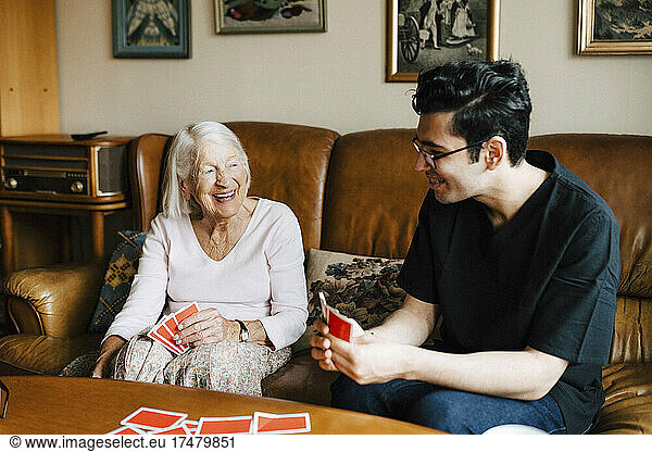 Happy male healthcare worker playing cards with elderly woman in living room