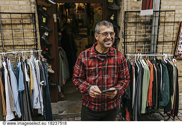 Happy male owner with mobile phone standing outside clothing store