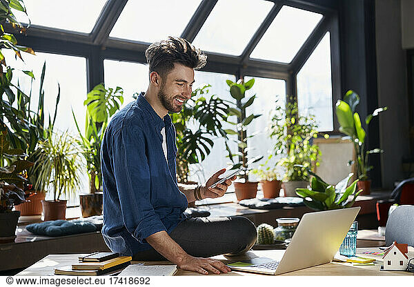 Happy male professional holding smart phone while sitting with laptop at desk in office
