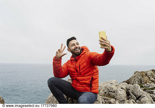 Happy man gesturing peace sign taking selfie through smart phone sitting on rock