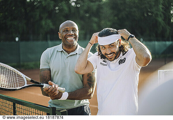 Happy man holding tennis racket with male friend adjusting headband at sports court
