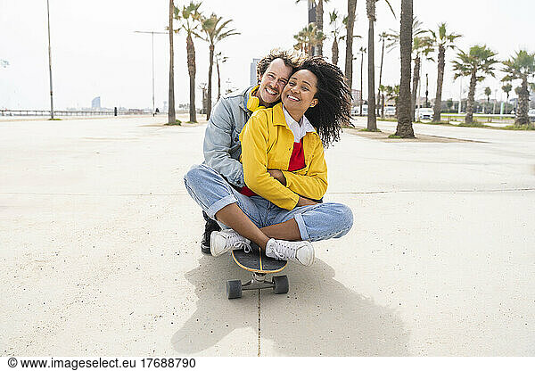 Happy man hugging woman sitting on skateboard in front of trees