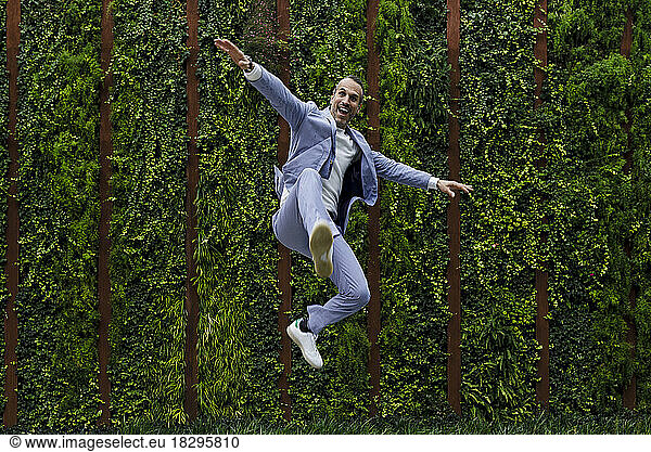Happy man jumping with arms outstretched in front of hedge