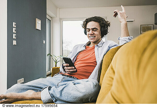 Happy man listening music through wireless headphones using smart phone on sofa at home