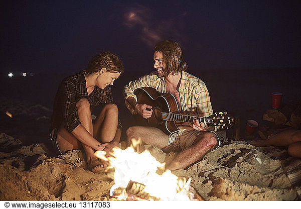 Happy man playing guitar for woman by bonfire at beach