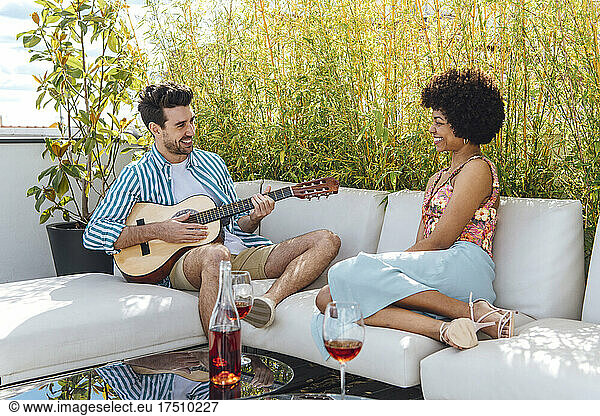 Happy man playing guitar for woman while sitting on sofa at penthouse patio