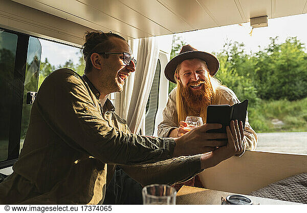 Happy man sharing smart phone with male friend in camping van