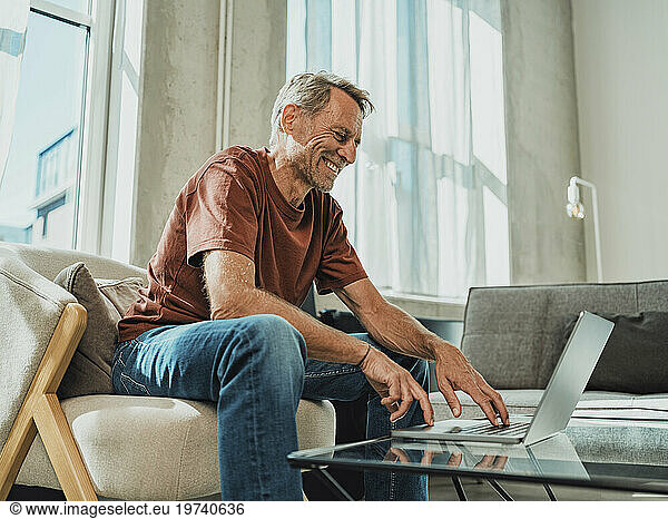 Happy man sitting on armchair and using laptop at home