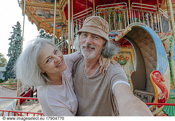 Happy man taking selfie with woman in front of carousel