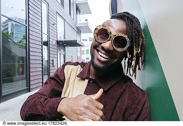 Happy man wearing sunglasses making thumbs up gesture leaning on wall