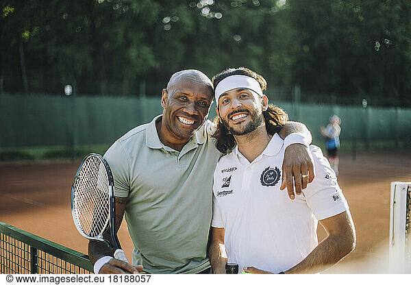 Happy man with arm around male friend wearing headband at tennis court