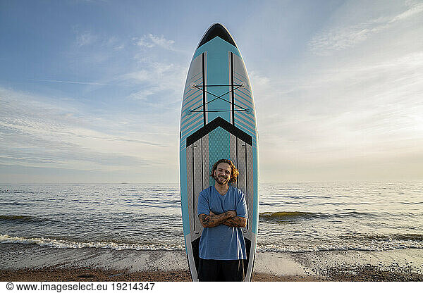 Happy man with arms crossed standing in front of paddleboard at beach