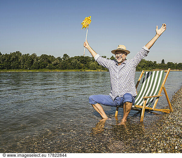 Happy man with arms outstretched holding paper pinwheel toy at riverbank