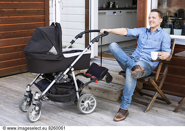 Happy man with baby carriage holding coffee mug while sitting at patio