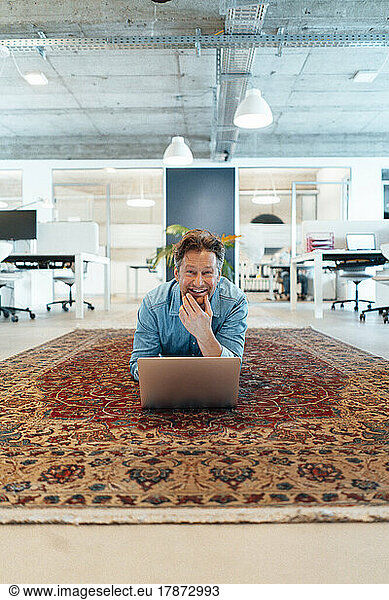 Happy mature businessman lying down with laptop on carpet in office