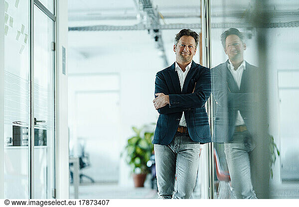 Happy mature businessman standing with arms crossed leaning on glass wall in office