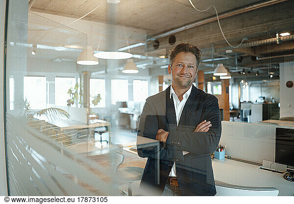 Happy mature businessman standing with arms crossed seen through glass in office