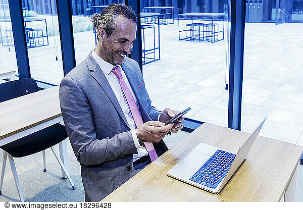 Happy mature businessman using smart phone by laptop at desk in office