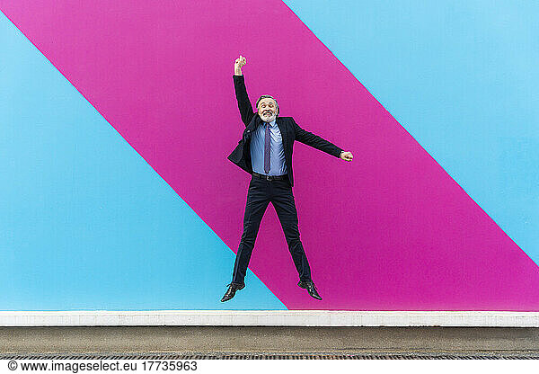 Happy mature businessman with hand raised jumping in front of pink and blue wall