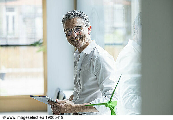 Happy mature businessman with tablet PC in front of window in office