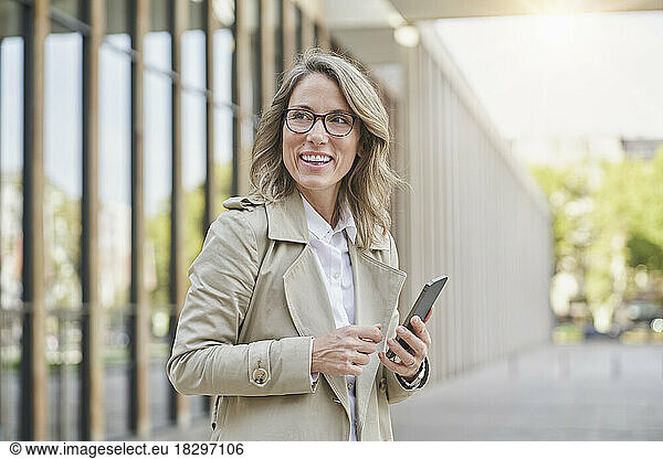 Happy mature businesswoman holding smart phone outside building