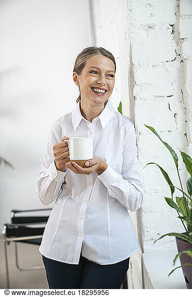 Happy mature businesswoman with coffee cup standing by wall
