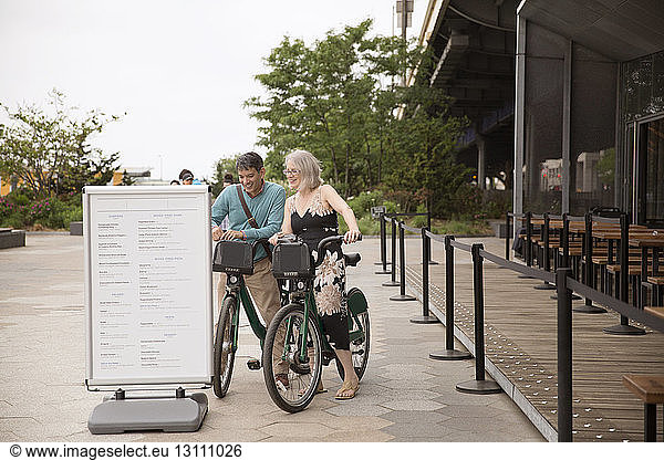 Happy mature couple reading information sign while standing with bicycles on street