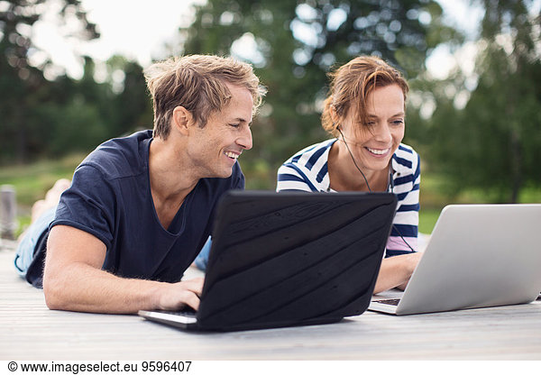 Happy mature couple using laptops while lying on pier