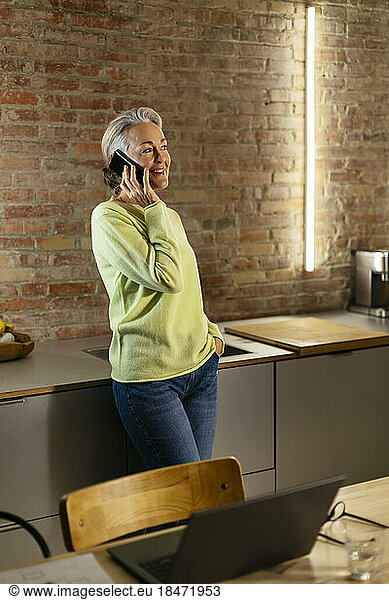 Happy mature freelancer talking on smart phone by kitchen counter