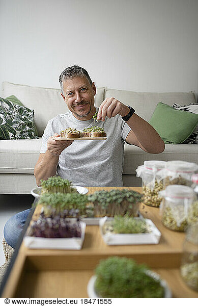 Happy mature man putting bean sprouts on toasted bread in living room at home