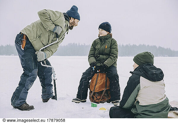Happy mature man using ice auger while talking with son sitting on backpack at frozen lake