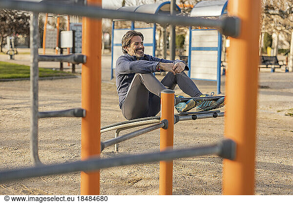 Happy mature man with hands clasped sitting on exercise equipment in park
