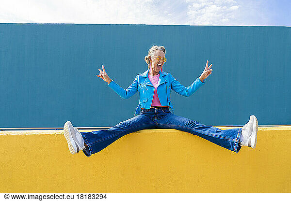 Happy mature woman gesturing peace on yellow wall
