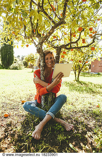 Happy mature woman hugging orange fruit tree and using tablet PC